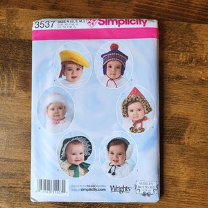 May include: A Simplicity sewing pattern for making six different baby hats. The pattern includes a yellow hat, a red and white striped hat, a white hat with a pom-pom, a brown and white hat with a fur trim, a white hat with a lace trim, and a gray hat with ear flaps. The pattern is for sizes XS, S, M, and L.