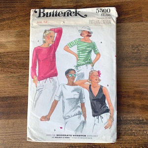 May include: A vintage Butterick sewing pattern envelope, size 12, featuring illustrations of women's tops. The designs include a long-sleeved pink top, a green and white striped top, a white top, and a black tank top. The envelope has the number 5500.