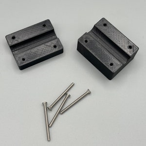 May include: Two black 3D printed brackets with four silver screws. The brackets have a slot in the centre and four holes for screws.