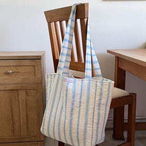Puede incluir: Un bolso tote acolchado grande con rayas verticales azules y blancas cuelga de una silla de madera. El bolso tiene correas largas y una pequeña etiqueta. La silla es de madera y está colocada frente a una pared de color claro.