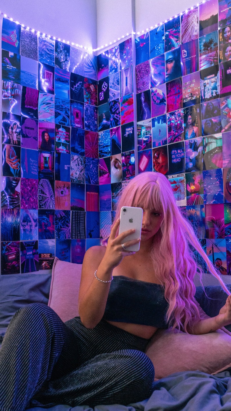 May include: A person with pink hair is sitting on a bed and taking a selfie with a white iPhone. The wall behind them is covered in a collage of colorful photos and images. The person is wearing a black crop top and striped pants.