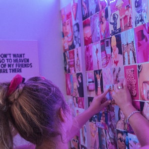 May include: A person adds a pink and white photo to a wall collage of various photos. The collage is made up of many different images, including lips, faces, and other abstract designs. The wall is decorated with white string lights.