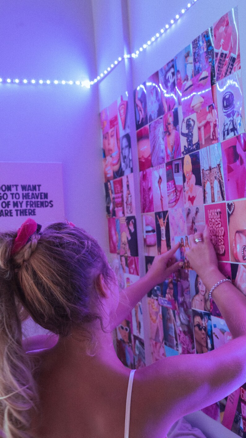May include: A person adds a pink and white photo to a wall collage of various photos. The collage is made up of many different images, including lips, faces, and other abstract designs. The wall is decorated with white string lights.
