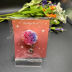May include: A badge reel with a colorful d20 die design. The reel features a pink, purple, and blue color scheme with white numbers. The card reads "Badge Reel" and "LUPUSNEUS.COM".