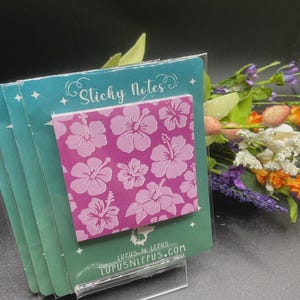 May include: A stack of sticky notes with a pink hibiscus flower pattern. The notes are displayed in a clear plastic holder. The packaging has the text "Sticky Notes" and the website address "LUPUSNLEPUS.COM".