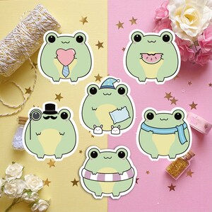 May include: Six different frog stickers with various expressions and accessories. The frogs are green with white eyes and are wearing items like a top hat, a scarf, a sleep mask, a heart, and a watermelon slice.