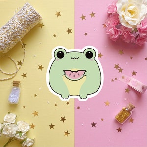May include: A green frog cartoon sticker with a cute expression, holding a slice of watermelon. The sticker is on a pink and yellow background with gold stars.