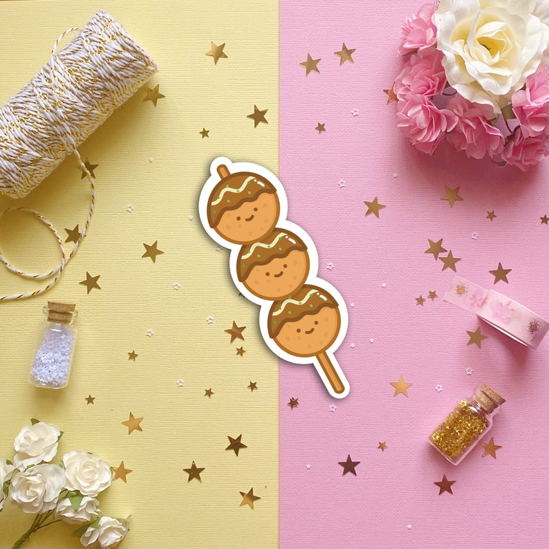 May include: A sticker of three brown takoyaki balls on a stick with smiling faces. The takoyaki are decorated with green onions and a brown sauce. The sticker is on a pink and yellow background with gold stars.