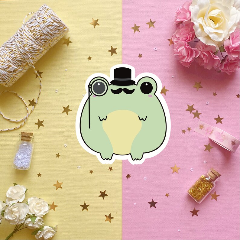 May include: A green frog wearing a black top hat and a monocle. The frog is sitting on a yellow and pink background with gold stars.