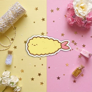 May include: A yellow cartoon shrimp tempura with a pink tail, surrounded by gold stars on a yellow and pink background.