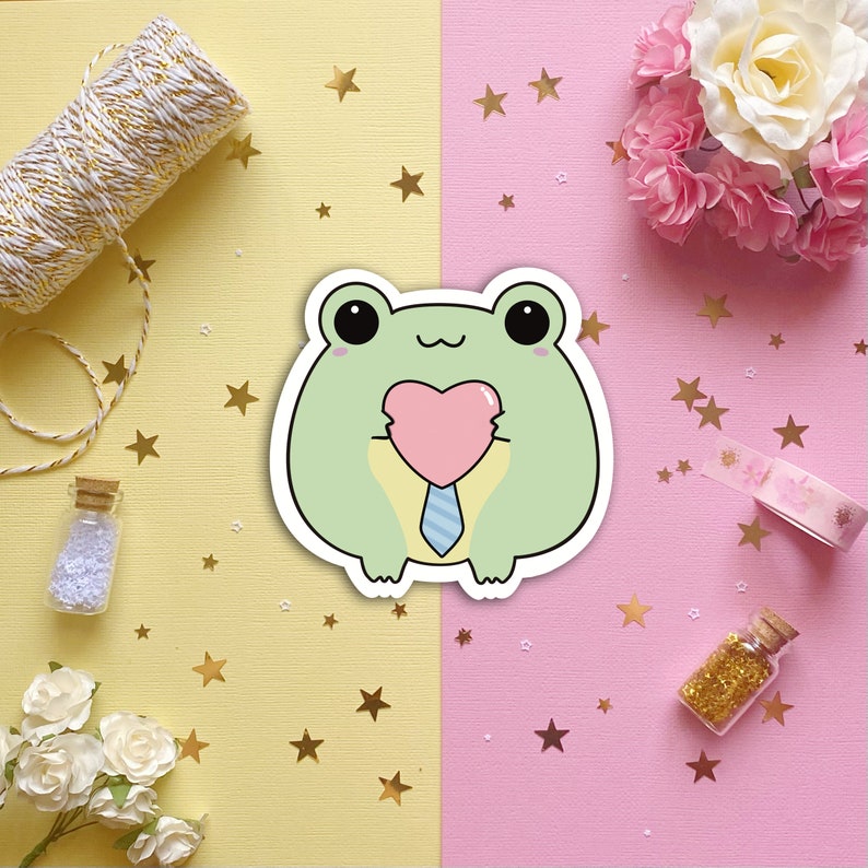 May include: A green frog cartoon sticker with a pink heart and a blue tie. The frog is holding the heart in its hands. The sticker is on a pink and yellow background with gold stars.