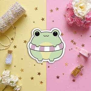 May include: A green frog wearing a pink and white striped life preserver. The frog is cartoon-style and has large eyes. The background is a split image of yellow and pink with gold stars.