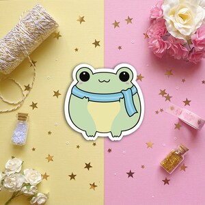May include: A green frog wearing a blue scarf, with a cute cartoon style. The frog is on a white background with a pink and yellow background behind it.