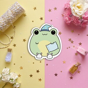 May include: A green frog wearing a blue and white nightcap holds a blue pillow and is surrounded by gold stars on a pink and yellow background.
