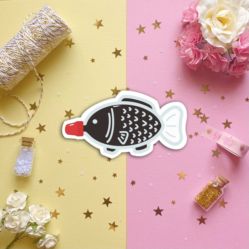 May include: A black and white fish-shaped sticker with a red cap on the front. The sticker is on a pink and yellow background with gold stars.