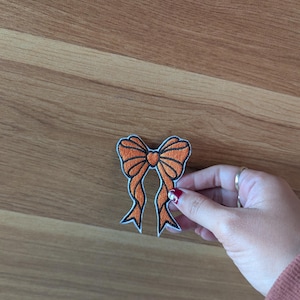 May include: Orange embroidered iron-on patch of a bow with a heart in the centre.