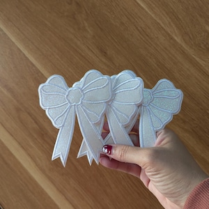 May include: Four white glitter bow patches with a satin finish. The bows have a center flower detail and are arranged in a stack.