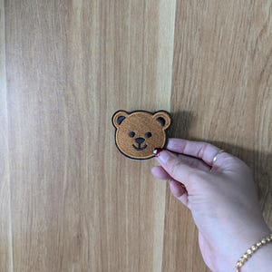 May include: A brown teddy bear patch with a black border. The bear has a black nose and eyes. The patch is held by a hand with a gold bracelet and a red painted fingernail.
