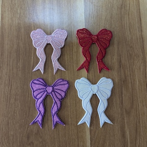 May include: Four glitter bow tie patches in pink, red, purple, and white. The bow ties are all the same size and shape, and they have a heart in the centre.