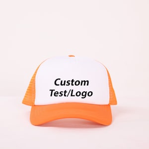 Op de afbeelding: Een witte en oranje truckercap met een mesh achterkant. De voorkant van de cap heeft de tekst "Custom Test/Logo" in zwart.