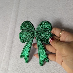 May include: A green glitter bow with a centre loop. The bow is made of fabric and has a shiny, sparkly finish.