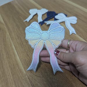 May include: A selection of decorative bow patches in various colours. The main bow, held by a hand, has a pastel gradient with glitter accents. Additional bows in white and navy blue are also visible.