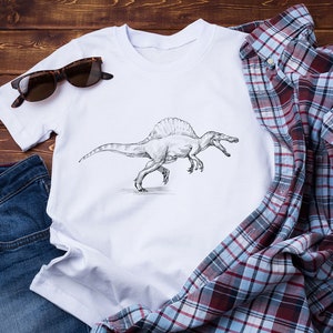 May include: A white t-shirt with a black and white illustration of a Spinosaurus dinosaur. The t-shirt is laid out on a wooden surface with a pair of sunglasses, blue jeans, and a plaid shirt.