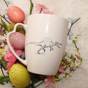 May include: White ceramic mug with a black and white illustration of a Spinosaurus dinosaur. The mug has a heart-shaped handle and is surrounded by pink and yellow Easter eggs and floral decorations.