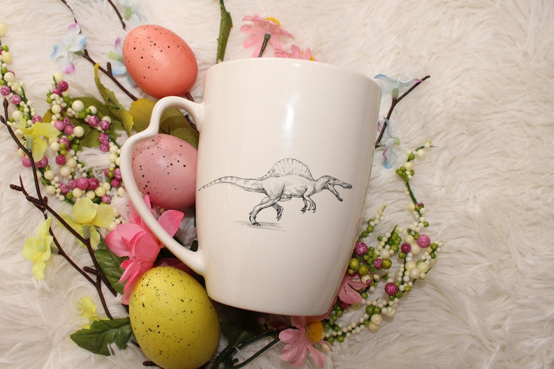 May include: White ceramic mug with a black and white illustration of a Spinosaurus dinosaur. The mug has a heart-shaped handle and is surrounded by pink and yellow Easter eggs and floral decorations.