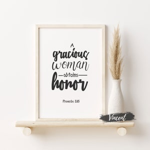 May include: A white framed print with black text that reads "A gracious woman obtains honor" and "Proverbs 11:16". The print is on a light wood shelf with a vase of dried grass.