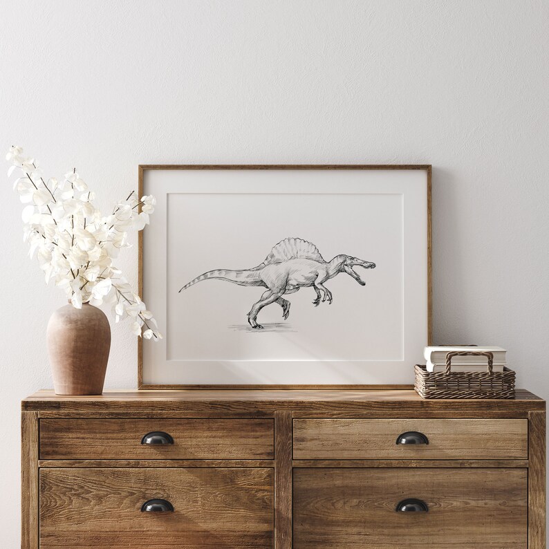 May include: A framed black and white pencil drawing of a Spinosaurus dinosaur, with a frill on its back, running with its mouth open. The drawing is on a white background and is framed in a light brown wood frame. The frame is sitting on a wooden dresser with two drawers.