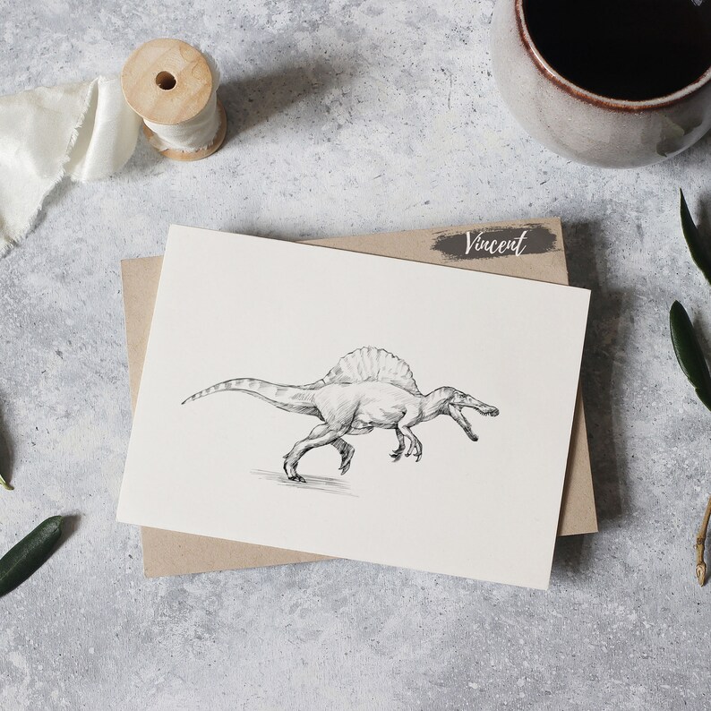 May include: A black and white drawing of a Spinosaurus dinosaur on a white card with the name "Vincent" written in black on a brown background.