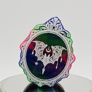 May include: A colorful, decorative bat-shaped pendant with a white bat silhouette inside a circular frame. The frame has an ornate, scalloped edge with pink, green, and blue hues. The background is a gradient of blue, green, and red.