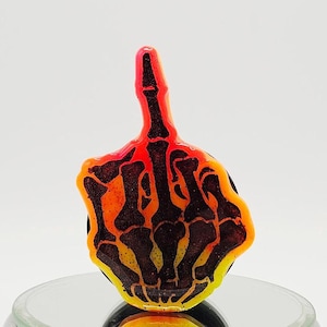 May include: A resin pop socket featuring a hand giving the middle finger. The design is a skeleton hand with a gradient of orange, yellow, and black. The pop socket is on a reflective surface.