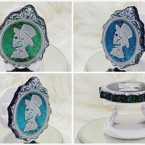 Op de afbeelding: Een witte telefoonhouder met een rond, zilveren frame en een groen-blauw, iriserend ontwerp. Het ontwerp toont een skelet dat een hoge hoed en een strikdas draagt.