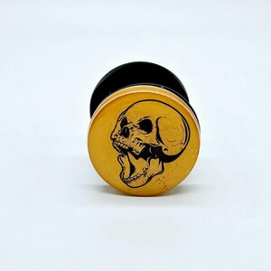 May include: A gold-coloured phone grip with a black skull design. The skull is open-mouthed and has a detailed design.