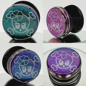 May include: Three pairs of iridescent ear gauges featuring a skull and crossbones design. The gauges are in shades of green, purple and blue.