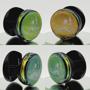 May include: Two pairs of skull-shaped glass plugs with black rubber backing. The glass plugs are green and yellow with a white skull design.