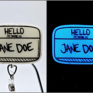 May include: A white name tag with black text that reads "HELLO MY NAME IS JANE DOE". The name tag is attached to a silver retractable badge holder. The name tag glows blue in the dark.
