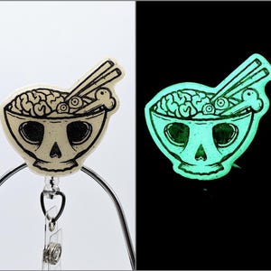 May include: A white glow-in-the-dark badge reel featuring a black and white illustration of a skull-shaped bowl filled with noodles, eyeballs, and bones. The reel is attached to a silver metal clip.
