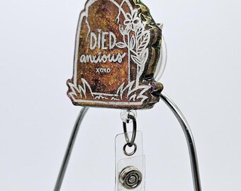 May include: A gold and green glitter resin tombstone with the text "DIED anxious xoxo" in white. The tombstone is attached to a clear plastic badge holder with a silver metal clip.
