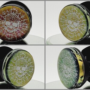 May include: Four different coloured glass skull plugs with a white skull and spiderweb design. The plugs are set against a black background.