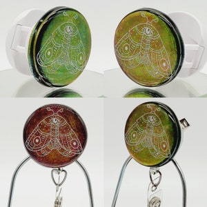 May include: A retractable badge holder with a green and gold iridescent background featuring a white line drawing of a moth with a single eye. The moth has a detailed design with intricate patterns on its wings.