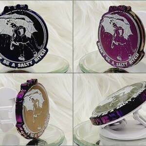 May include: A round phone grip with a design featuring a person under an umbrella in the rain, with the text "DON'T BE A SALTY BITCH". The grip has a white base and a metallic purple and black rim.