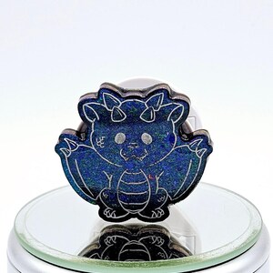 May include: A small, blue, iridescent dragon figurine with a white outline. The dragon has a cute, cartoon-like expression and is sitting on a round, silver base.
