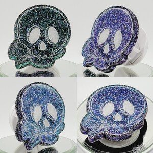 May include: Four phone grips with a skull design. Each grip has a different color scheme with glitter accents. The skulls are outlined in black and have a floral design around them.