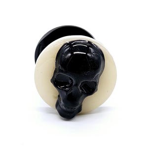 May include: A black skull design on a white circular plug with a black backing.