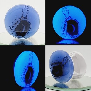 Könnte beinhalten: Ein blauer Glasflaschenverschluss mit einer schwarz-weißen Illustration einer Flasche mit einem Etikett, auf dem "Dream" steht. Der Flaschenverschluss leuchtet unter Schwarzlicht blau.