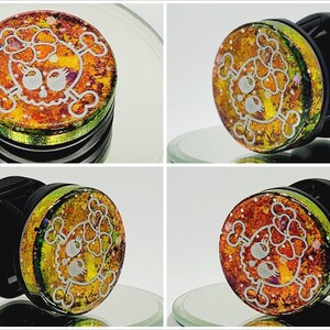 May include: A pair of black and green ear gauges featuring a skull and crossbones design. The skull has a bow on top and is set against a glittery, iridescent background.