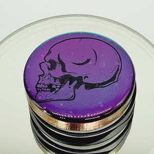 May include: A purple and black skull design on a round, iridescent sticker. The sticker is on top of a stack of black and gold rings.